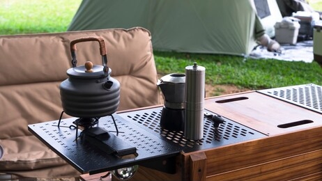 vintage outdoor black kettle and coffee grinder on camping table with armchair on green lawn in camping area