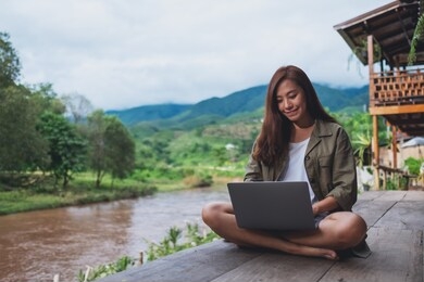 portrait image of a beautiful asian woman working on laptop computer while sitting by the river with mountains and nature background