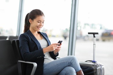 airport business woman on smart phone at gate waiting in terminal. air travel concept with young casual businesswoman sitting with hand luggage suitcase. beautiful young mixed race female professional