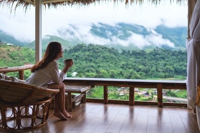 portrait image of a beautiful asian woman holding and drinking hot coffee , sitting on balcony and looking at mountains and green nature 