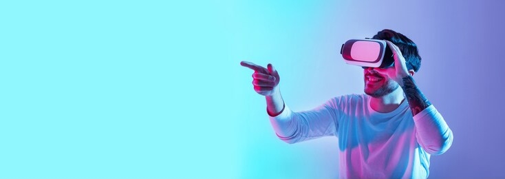 acquaintance with virtual world. smiling guy with glasses points finger in neon on blue and violet background, panorama, copy space