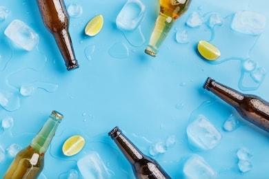 summer party and chilled drinks. dark and light glass beer bottles without labels with ice and pieces of lime on blue background, top view, free space