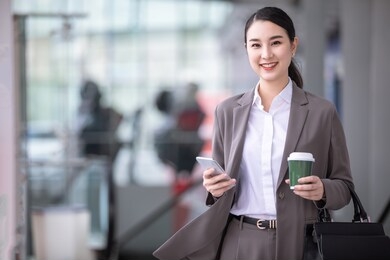 asian woman with smartphone standing against street blurred building background. fashion business photo of beautiful girl in casual suite with phone and cup of coffee