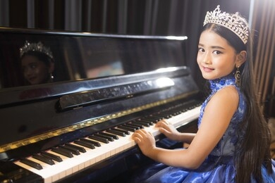 a portrait of a cute asian girl, makeup, wearing a blue evening dress and a crown, playing the piano in the room.