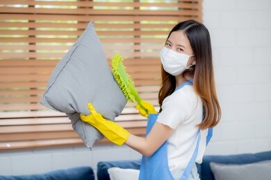 young asian woman in face mask and gloves cleaning dust with duster on sofa and cushions at home, girl doing housework for hygiene at house, housekeeper and service, health care and lifestyle concept.