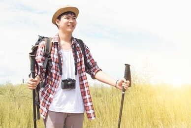 an asian handsome man with backpack wearing hat and check shirt, is hiking and standing in green field with background of blue sky. summer and travel concept.