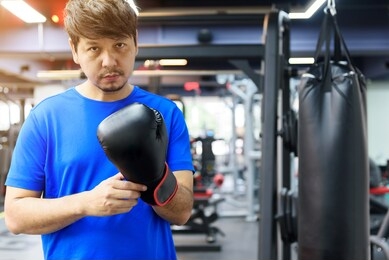 handsome asian man wears a blue sport shirt with black boxing gloves in the gym look at camera, there is sandbag on the background, sport and workout concept