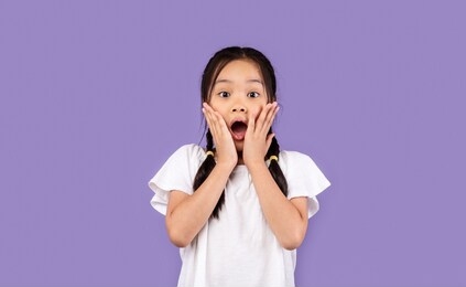 omg. shocked asian kid girl touching face looking at camera in amazement standing over purple background. studio shot