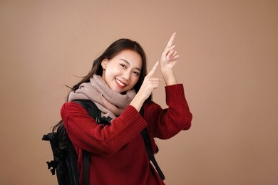 asian tourist woman backpacker point to the top, winter outfit wear red sleeves and pink scarf, isolated mocha brown background