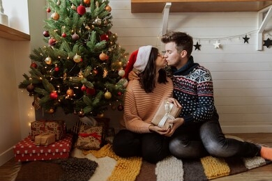 stylish happy couple exchanging christmas gifts under christmas tree with lights. young family kissing and holding present, happy moment in festive modern room. merry christmas