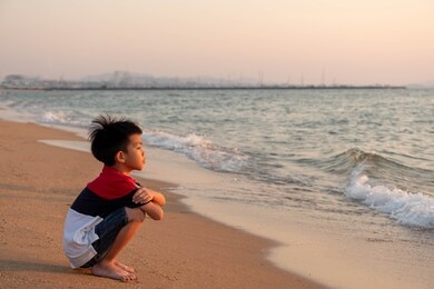cute toddler asian boy sitting alone on the beach at sunset. child sitting on the beach near sea and looking on the waves, silhouette of the boy on the seaside in sunset.