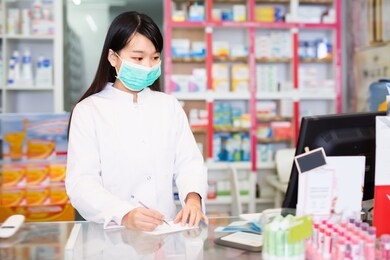 adult chinese female druggist in protective facial mask working in pharmacy