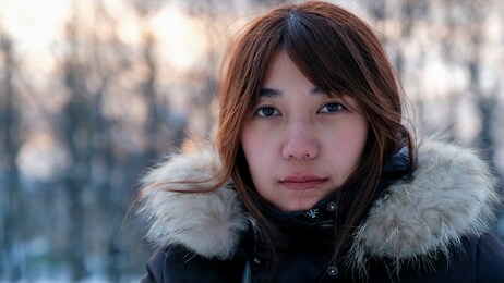 portrait of young asian woman happy in warm black winter suit with light warm fur with blur forest background and some orange lighting in twilight. concept of girl traveling in winter outfit.