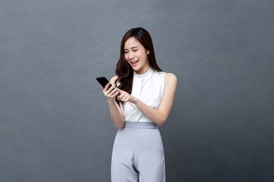 portrait of smiling young asian businesswoman holding and texting on mobile phone in isolated studio gray background