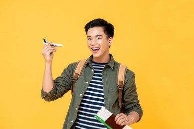 fun travel concept portrait of smiling young male asian tourist holding airplane toy with passport and ticket in isolated studio yellow background