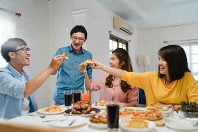 happy young friends group having lunch at home. asia family party eating pizza food and laughing enjoying meal while sitting at dining table together at house. celebration holiday and togetherness.