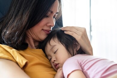 asian beautiful mother hugging sleeping baby in her arms and kissing the kid gently. the mom closing her eyes while holding her baby head to rest on shoulder. touch of love and family relationship.