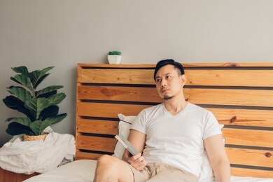 asian man is reading book on his bed in late afternoon.