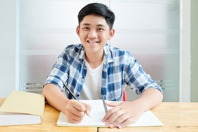 portrait of smiling asian schoolboy writing and drawing in copybook when sitting at desk