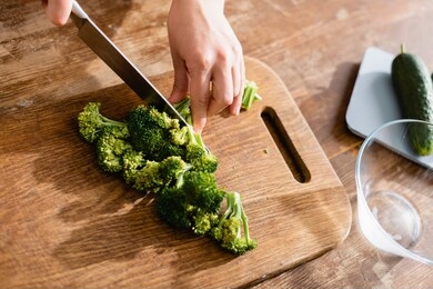 partial view of woman cutting fresh broccoli on chopping board near kitchen scales with cucumbers