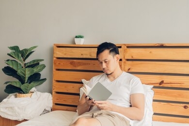 asian man is reading book on his bed in late afternoon.
