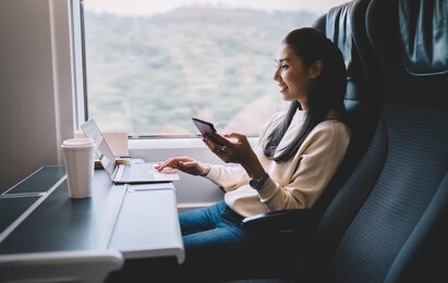 side view of positive ethnic female in casual outfit with smartphone working on laptop and smiling while resting on comfortable armchair near window of train