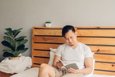 asian man is reading book on his bed in late afternoon.