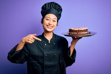 young chinese chef woman wearing cooker uniform and hat holding tray with cake with surprise face pointing finger to himself