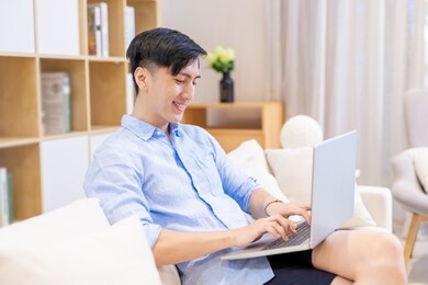 asian young man is sitting on the sofa and using laptop leisurely at home