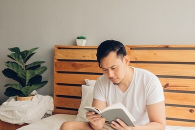 asian man is reading book on his bed in late afternoon.