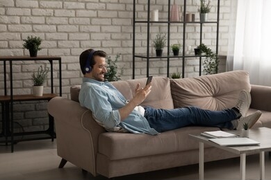 happy young caucasian man in earphones sit relax on couch in living room browse internet on modern cellphone gadget, smiling millennial male in headphones listen music on smartphone at home