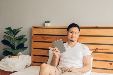 asian man is reading book on his bed in late afternoon.