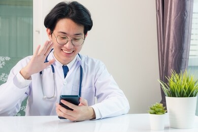 asian doctor young handsome man smiling video call raise hands to greet patients with digital smart mobile phone at hospital desk office, technology healthcare medical concept
