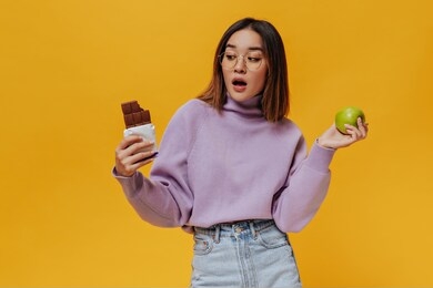 surprised shirt-haired girl in eyeglasses looks at milk chocolate bar. charming asian woman in purple sweater and denim skirt holds green fresh apple on orange background.