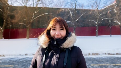 woman traveler asian female in warm black winter outfit with light brown fur smile under sunlight with snow and red wall as background. thai lady tourist in cold country.