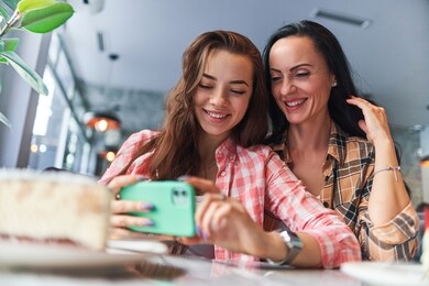 happy smiling mother and joyful teen daughter watch video content on the phone and have good time together in a cafe