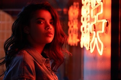 attractive african american woman standing lit with red neon light near sign in night club. stylish cool fashion young mixed race hipster looking at camera at 80s 90s gen z retro style party.