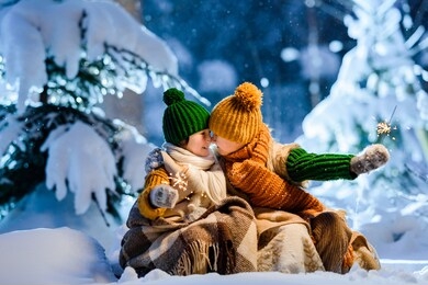 two little kids celebrating christmas outdoors holding sparklers in hands. children wrapped themselves in warm woolen plaid. fairy winter night, snow-covered christmas trees around.