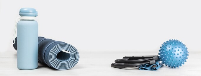 rolled blue yoga mat. exercise resistance band water bottle on grey wooden surface. gender neutral fitness and exercise concept. copy space. active lifestyle sport. workout at home or gym web banner