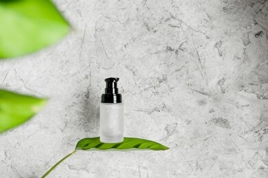 glass jar with gel and black lid-dispenser without inscriptions stands on green leaf of plant on gray concrete background, cosmetic layout