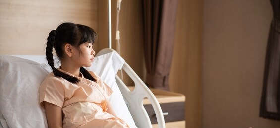sick asian little girl sitting on hospital bed feeling sad and unhappy. homesick girl looking hopefully outside and want to go home soon while being treated in the hospital. banner ratio photo.