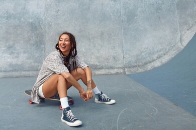 portrait of skater girl in skatepark. female teenager in casual outfit sitting on skateboard against concrete wall. summer skateboarding with modern sport equipment as part of active lifestyle.