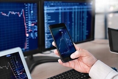 man trader sitting at desk at office in front of monitors with stocks data holding smartphone browsing application monitoring price changes on candle bar online using digitla devices close-up