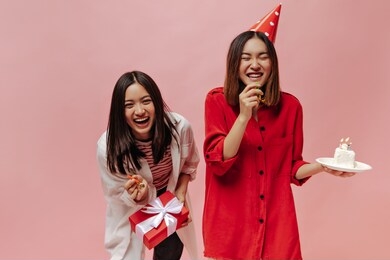 joyful brunette asian women laugh on pink isolated background. lady in party hat and red blouse holds birthday cake. girl in striped shirt poses with gift box.