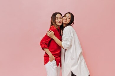 brunette asian woman in long oversized shirt and cheerful short-haired girl in red blouse and white pants hug on pink background.