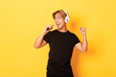 carefree handsome asian guy in headphones, playing karaoke app, singing into mobile phone microphone, standing yellow background