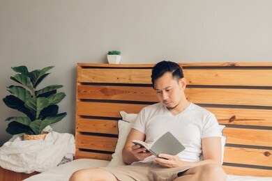 asian man is reading book on his bed in late afternoon.