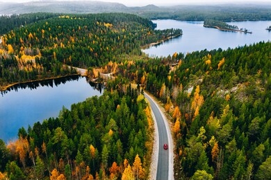 aerial view of rural road with red car in yellow and orange autumn forest with blue lake in finland.