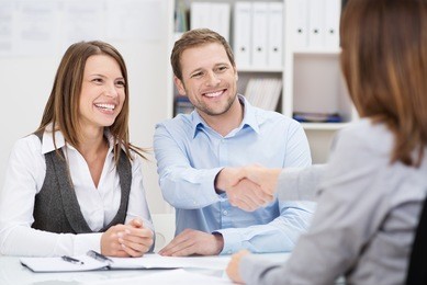 smiling young man shaking hands with an insurance agent or investment adviser as he sits in a meeting with his wife in her office