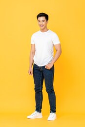 full length portrait of smiling young handsome asian man standing with one hand in pocket isolated on yellow studio background